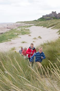Family of four running up the sand dunes at the beach. They are wearing warm casual clothing and a castle can be seen in the background.