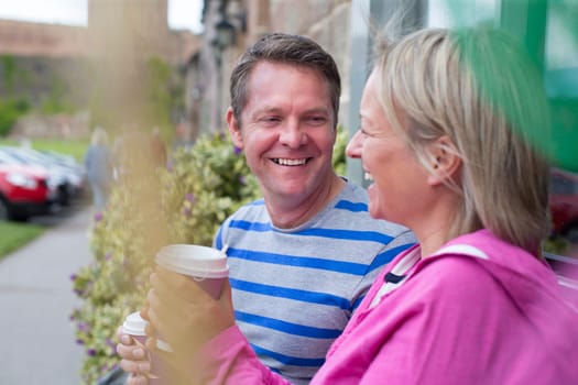 Mature couple laughing as they sit outside and drink hot drinks out of takeaway cups. They are wearing casual clothing and look very happy.