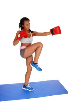 A slim African American girl standing on a blue mat during boxing exercise.with one leg up and red gloves, isolated for white background..