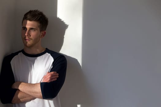 Portrait of an attractive young man, standing against a white wall with sunlight from the window shining on him.