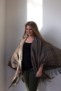 Portrait of an attractive young woman posing with a big scarf. She is standing in the sunlight and looking out the window.