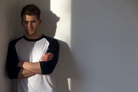 Portrait of an attractive young man, standing against a white wall with sunlight from the window shining on him.
