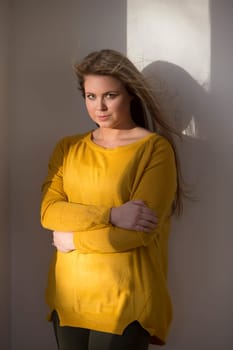 Portrait of an attractive young woman. She is standing in the light from the window and looking at the camera with her arms folded.