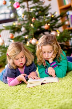 two blond girls reading a book in front of christmas tree