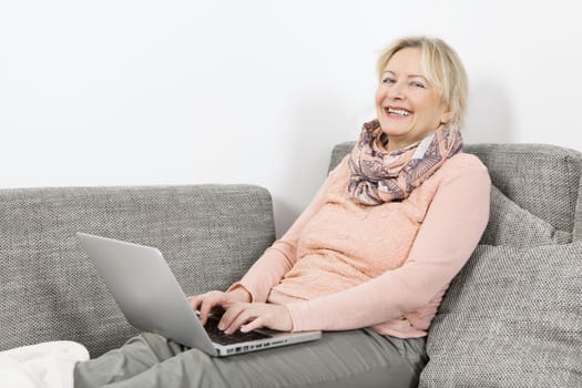 an older blonde woman lies on the sofa and works with a laptop