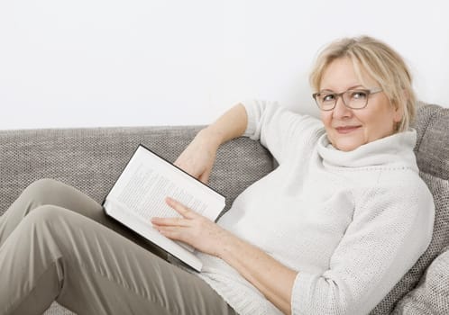 an older blonde woman lies on the sofa and reads a book