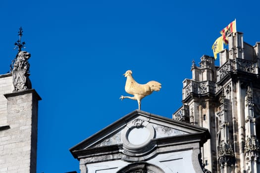 Facade of buildings at the Grote Markt (Large market square) in Mechelen, Belgium.  A golden rooster is irritating a lion neighbor . In the background  Flemish flags on the Rumbold tower of the Rumbold cathedral