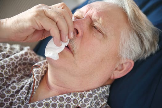 Sick senior man lies on sofa, and tends to his runny nose.