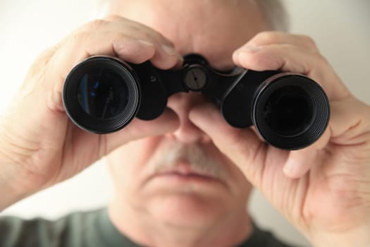 A senior man holds an old pair of binoculars pointed at the camera.