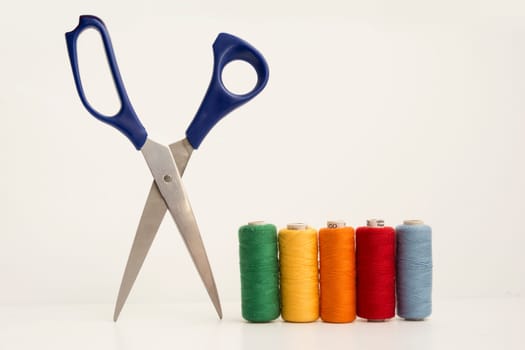 Multicolor Sewing Threads, Bobbins Beside Open Leged Blue Scissor Isolated on White Background.