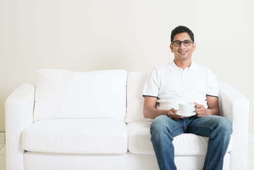 Young single Indian guy sitting on sofa alone and drinking coffee, copy space at side. Lifestyle Asian man at home. Handsome male model.