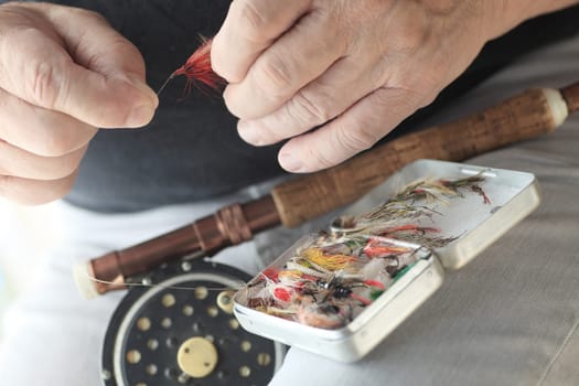 A fisherman with fly fishing pole and box of artificial lures holds a red fly.