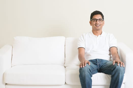 Young single Indian guy sitting lonely on sofa and smiling. Lifestyle Asian man at home. Handsome male model.