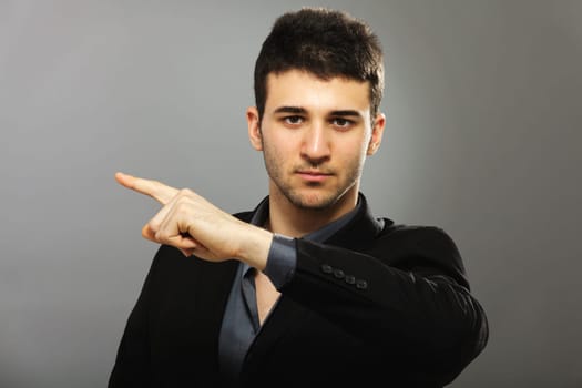 Young businessman looking at the camera and pointing with his left hand, isolated on gray