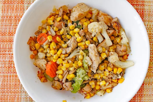 Cooked meal of chicken, carrots, broccoli, cauliflower and corn with pepper served in a round white bowl on a colorful placemat. Chicken and mixed vegetables supper.