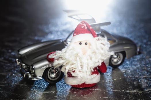 Santa Claus or Father Frost with old vintage automobile on on silver or metal grunge surface with backlight from behind. Main focus of image on Santa Claus and selective on turned left and opened doors toy retro car