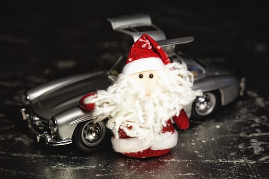 Santa Claus or Father Frost with old vintage automobile on on silver or metal grunge surface. Main focus of image on Santa Claus and selective on turned left and opened doors toy retro car
