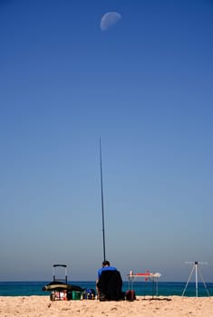 Sitting man fishing at the beach with the moon over blue sky, Tarifa, Spain