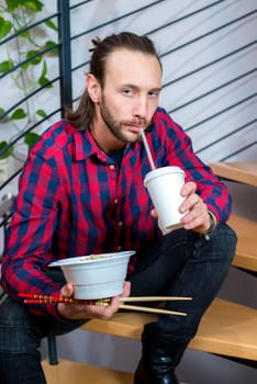 young man in checkered shirt sitting on stairs and eating asian food