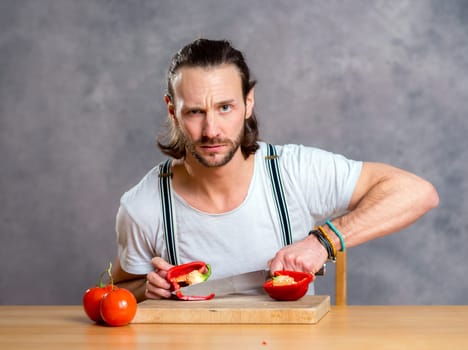 young bearded man cutting sweet pepper in front of gray background