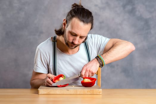 young bearded man cutting sweet pepper in front of gray background