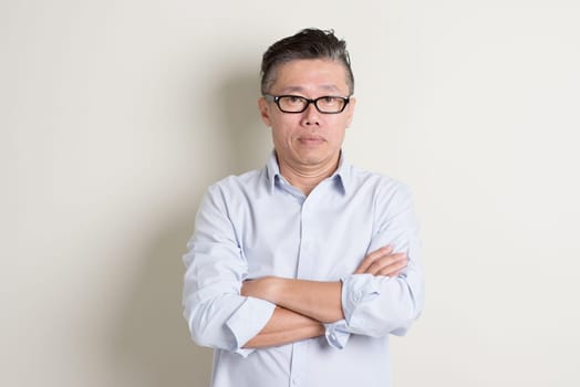 Portrait of single mature 50s Asian man in casual business arms crossed and standing over plain background with shadow.