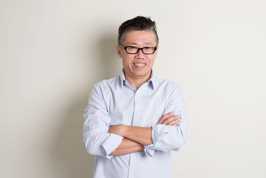 Portrait of confident single mature 50s Asian man in casual business arms crossed and standing over plain background with shadow.