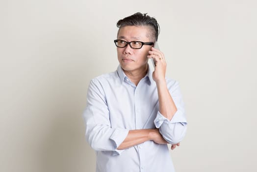 Portrait of single mature 50s Asian man in casual business calling on  smartphone, standing over plain background with shadow. Chinese senior male people.