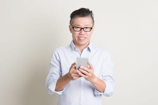 Portrait of single mature 50s Asian man in casual business using mobile applications on smartphone and smiling, standing over plain background with shadow. Chinese senior male people.