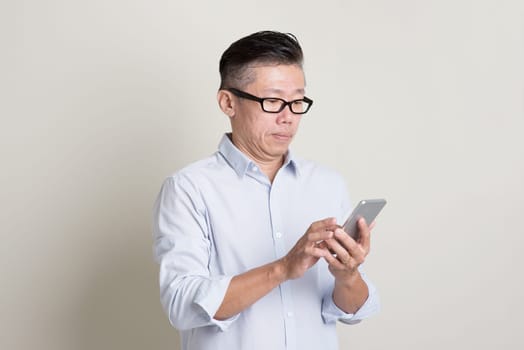 Portrait of single mature 50s Asian man in casual business playing smartphone, standing over plain background with shadow. Chinese senior male people.