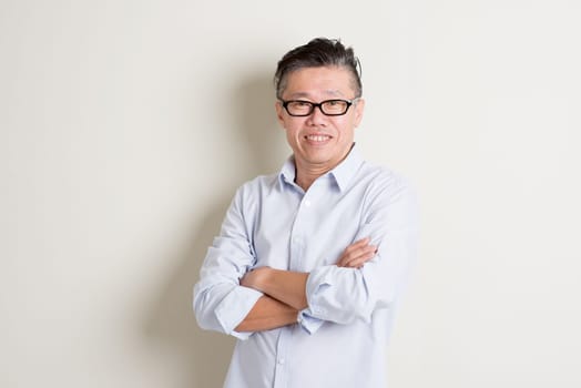 Portrait of happy single mature 50s Asian man in casual business arms crossed smiling and standing over plain background with shadow.
