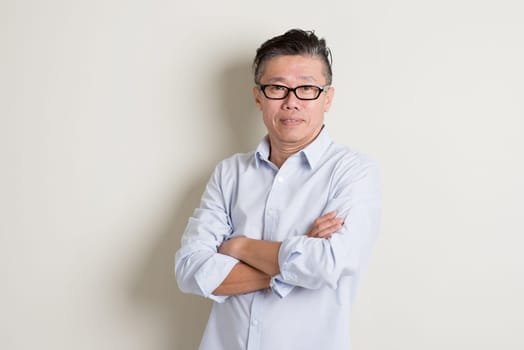 Portrait of single mature 50s Asian man in casual business arms crossed smiling and standing over plain background with shadow.