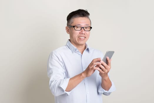 Portrait of single mature 50s Asian man in casual business texting using smartphone and smiling, standing over plain background with shadow. Chinese senior male people.