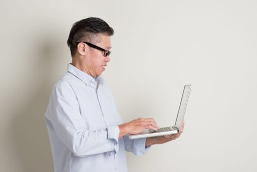 Portrait of single mature 50s Asian man in casual business using notebook computer and smiling, standing over plain background with shadow. Chinese senior male people.