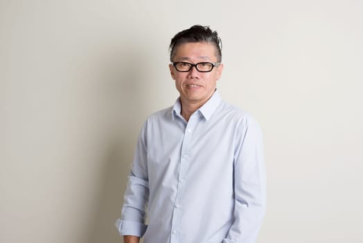 Portrait of single mature 50s Asian man in casual business smiling and standing over plain background with shadow.