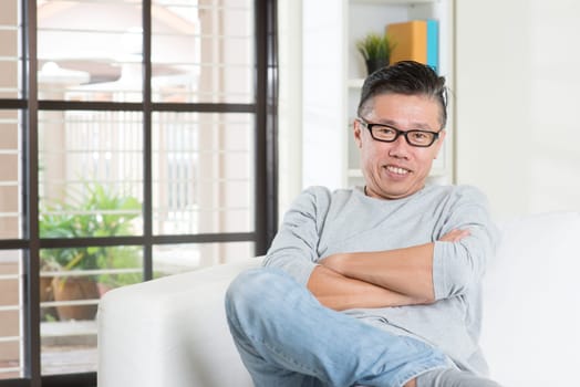 Portrait of happy mature 50s Asian man smiling and sitting at home. Senior Chinese male relaxed and seated on sofa indoor.