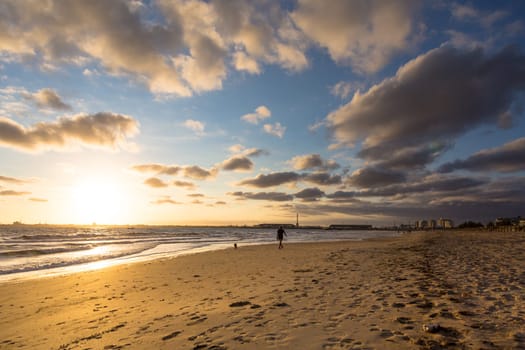 Sun setting on an urban beach on a cloudly afternoon, sand in the foreground with a person walking their dog.