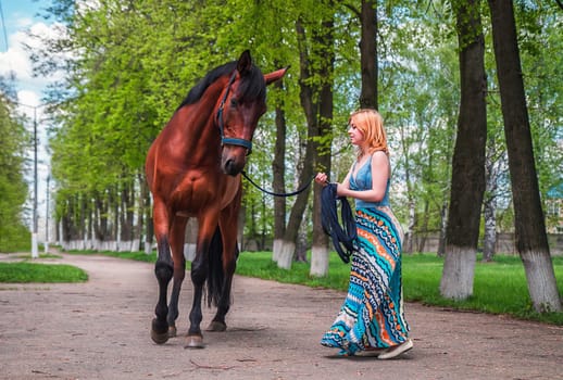 A young blond woman in a long blue dress leads the brown horse on the track along the high tree