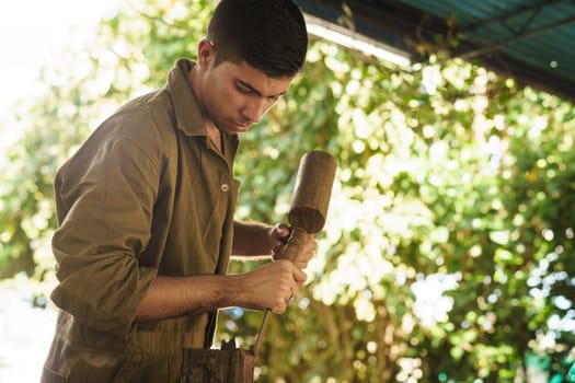 Young man at work learning craftsman profession, working with hammer and ceasel. The artist carves a raw block of wood to make a wooden statue