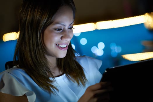 Beautiful hispanic girl typing on touch screen device, sitting at home at night. The young woman is studying on her tablet, then smiles looking at the screen