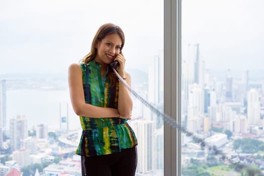 Young Hispanic woman leaning on table in a modern office building, with a beautiful sight of the city in background. She holds a wired telephone with long wire and talks looking away. Copy space