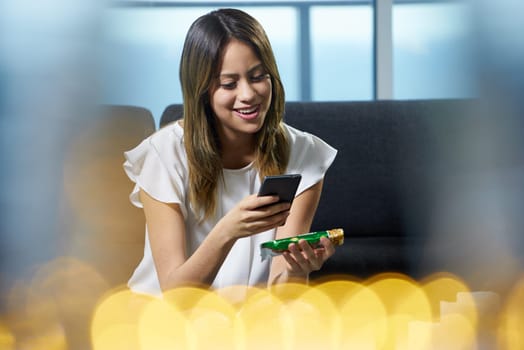 Young hispanic woman at home, holding a cereal bar and reading nutritional facts on package. She uses an app on her phone to scan the label and gets more information on calories and ingredients