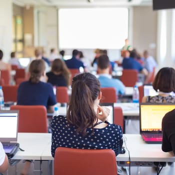 Workshop at university. Rear view of students sitting and listening in lecture hall doing practical tasks on their laptops. Square composition.