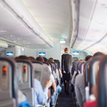 Interior of commercial airplane with passengers on seats during flight. Stewardess in dark blue uniform walking the aisle. Square composition.