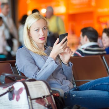 Casual blond young woman using her cell phone while waiting to board a plane at the departure gates. Wireless network hotspot enabling people to access internet conection. Public transport.