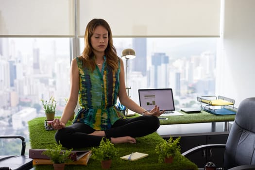 Young hispanic woman in office, sitting on desk covered with grass and plants. The businesswoman does zen and yoga meditation in lotus position. Full length, front view