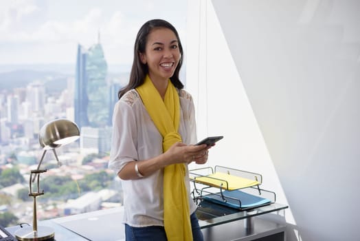 Young multiethnic Chinese Hispanic woman leaning on table in a modern office building, with a beautiful sight of the city in background. She holds a mobile phone and smiles at camera