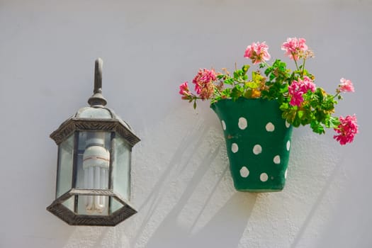 Green dotted flowerpot and pink flowers on a white wall with vintage lantern. Andalusian town, Spain