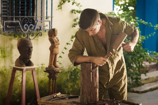 Young man at work learning craftsman profession, working with hammer and chisel. The artist carves a raw block of wood to make a wooden statue