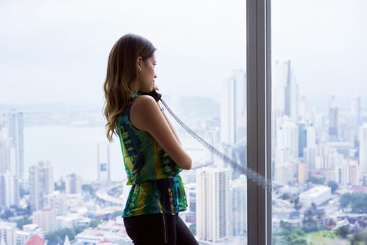 Young hispanic businesswoman in modern office building, with a beautiful sight of the city. She holds a wired telephone with long wire and looks outside window